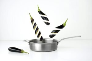 Creative image of sliced eggplants floating above a stainless steel pan against a white background.