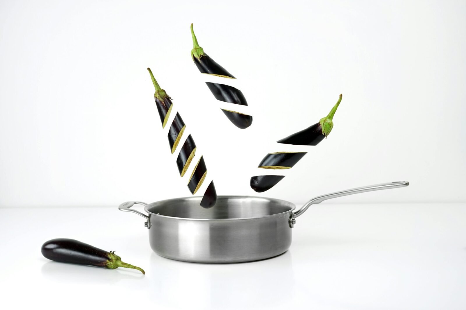 Creative image of sliced eggplants floating above a stainless steel pan against a white background.